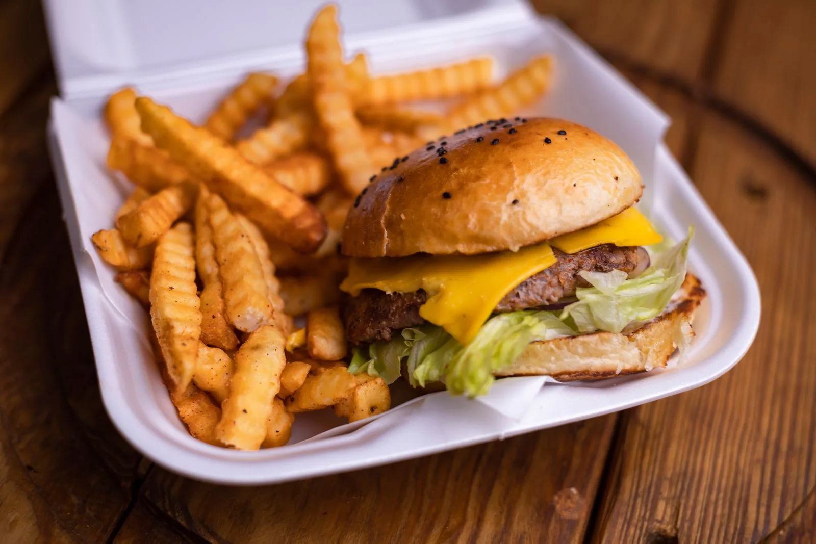 Cheeseburger and crinkle-cut fries in an open takeout box on a wooden table